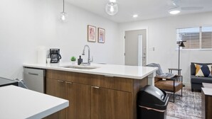 Una cocina moderna con encimera blanca, armarios de madera, cafetera y pileta. Hay una zona de comedor con una silla de cuero y una mesita.