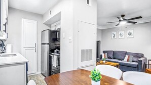 Een ruimtelijk ingerichte leefruimte met een kitchenette, een eethoek met een houten tafel, een grijze bank en een plafondventilator.