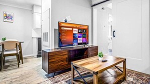 Een moderne woonkamer met een flatscreen-tv waarop streamingdiensten worden getoond, een houten salontafel, een eethoek met een stoel, en een keuken met een magnetron.