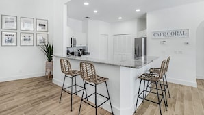 A modern kitchen with a marble island, black metal bar stools, and a potted plant. - Luxe Home, Pool, Boat Rental, Hot tub, & Fire pit (Cape Coral)