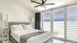 Quarto moderno com cama, ventilador de teto e vista do exterior por grandes janelas.