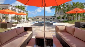 Uma área de piscina com duas grandes ombrelhas laranja, sofás de vime e uma piscina com revestimento de azulejos.