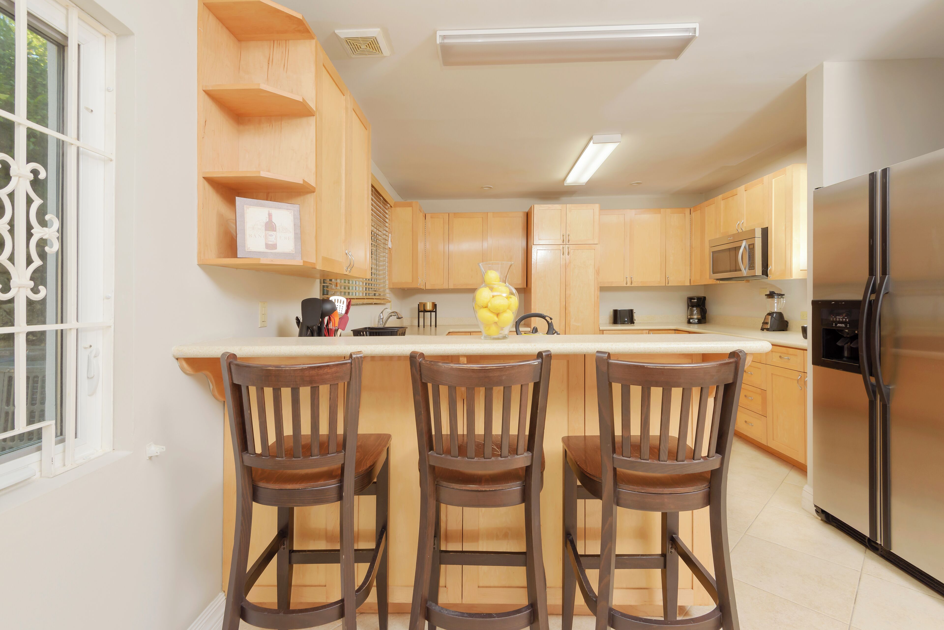 A kitchen with wooden cabinets, a stainless steel refrigerator, and a bar area with three wooden chairs.