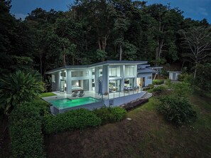 A modern, multi-story house with a large glass facade, a swimming pool, and a lush garden.