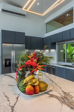 A modern kitchen with dark cabinets, a stainless steel refrigerator, a marble countertop, and a fruit bowl with tropical flowers.