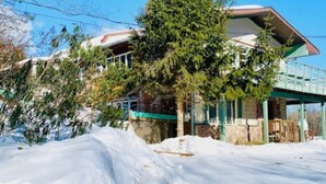 A two-story building with a snow-covered ground, a large evergreen tree in front, and a clear blue sky. - Catskill Cabin Hummingbird Suite 2 (Shandaken)