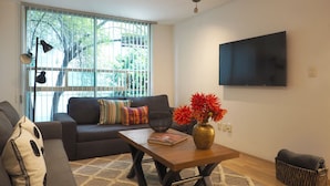 Una sala de estar con un sofá, una mesa de centro, un florero con flores, un televisor y una ventana con persianas.