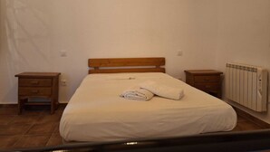 A bedroom with a wooden bed frame, a white mattress, and a rolled towel on the bed. There are two wooden nightstands, one on each side of the bed. A radiator is on the wall to the right of the bed.