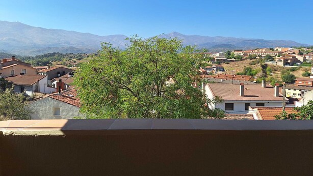 A view from a balcony with a wooden ceiling, overlooking a town with red-tiled roofs and a mountain range in the background.