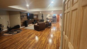 Amplia sala de estar con piso de madera, un sofá de cuero, un televisor montado en la pared y una cocina al fondo.