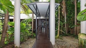 Un pasaje cubierto con estructura metálica y suelo de madera, rodeado de vegetación tropical.