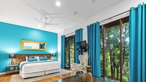 Un dormitorio con cama, una silla, una mesa decorada con una piña, un televisor y vistas a zonas verdes a través de amplias ventanas.