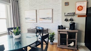 Zona de menjador moderna amb taula de vidre, cadires negres, una planta en test i racó de cafè amb cafetera i tasses, més obra d'art a la paret.