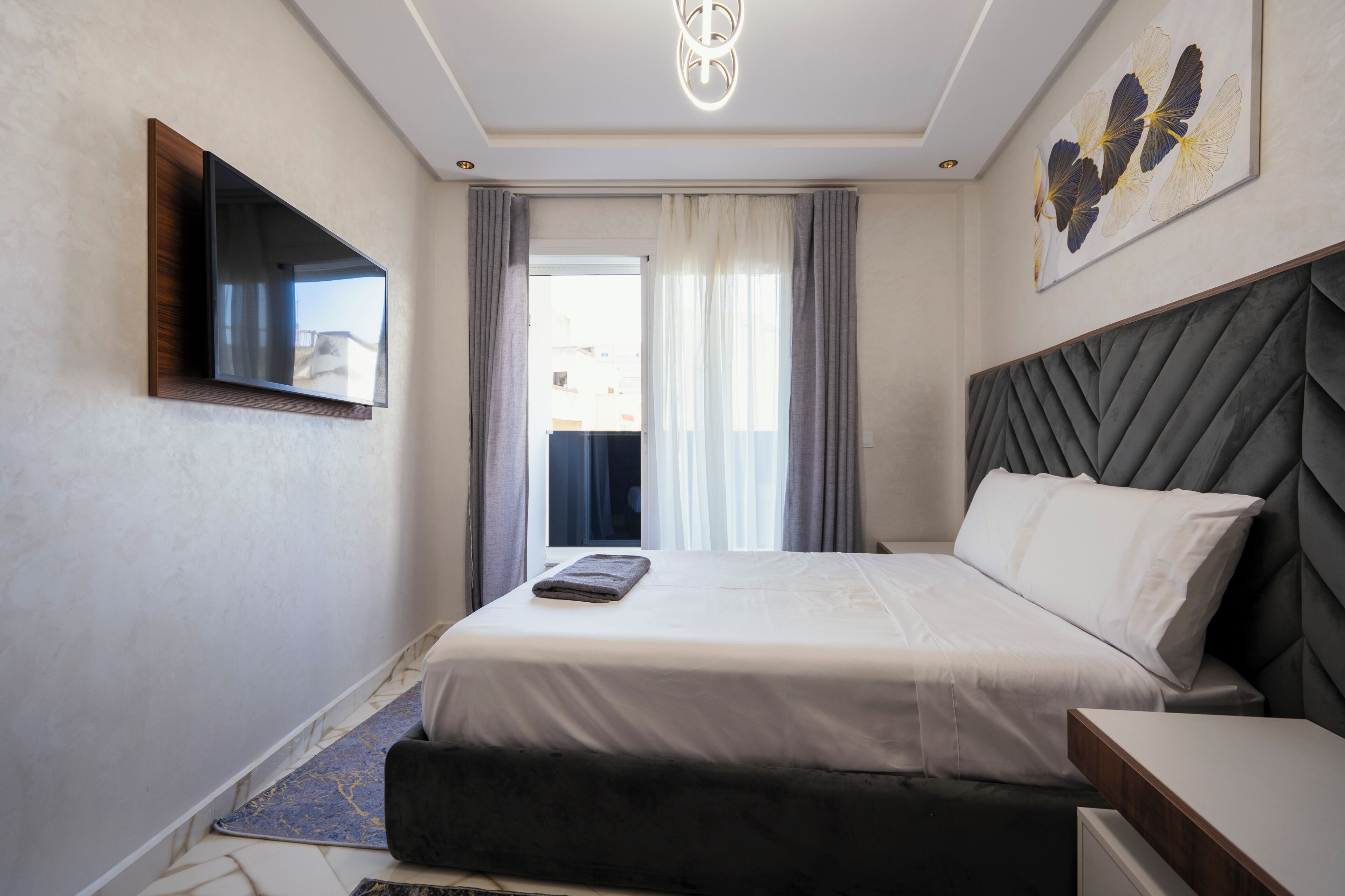 A modern hotel room with a bed, a flat-screen TV mounted on the wall, a window with curtains, and a decorative wall piece.