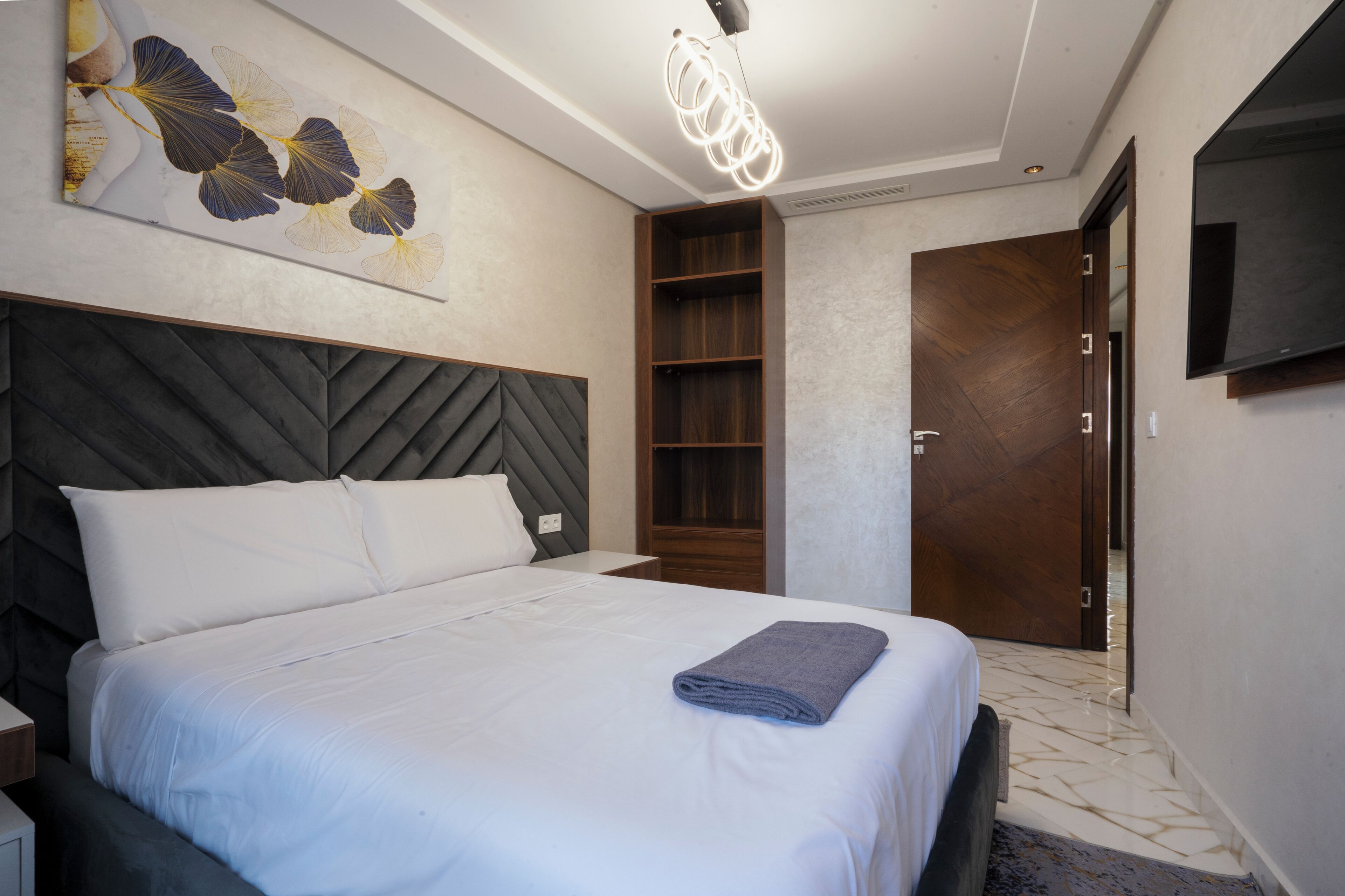 A modern hotel room with a large bed, a wooden headboard, a wall-mounted TV, and a built-in shelf.