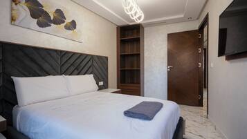 A modern hotel room with a large bed, a wooden headboard, a wall-mounted TV, and a built-in shelf.