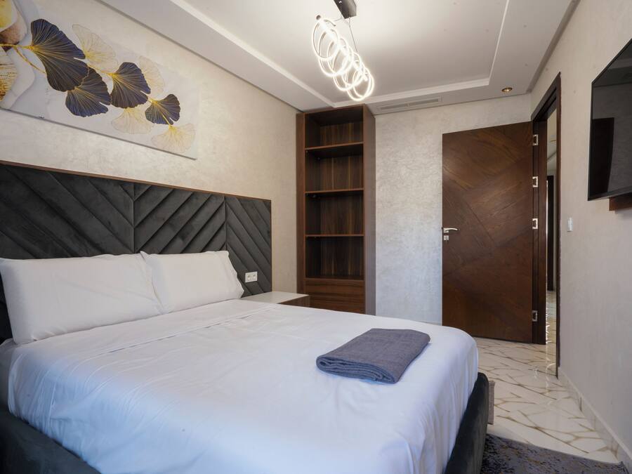 A modern hotel room with a large bed, a wooden headboard, a wall-mounted TV, and a built-in shelf.