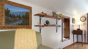 Ein Wohnzimmer mit einem grünen Sofa, einem Landschaftsbild, einem Holzregal mit Büchern und einer kleinen Figur, einer Wanduhr und einer Lampe.