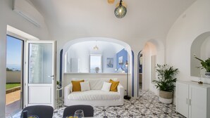 Une pièce avec un canapé blanc, une table à manger avec des assiettes et des verres, une plante en pot et une vue sur l’extérieur, visible à travers une porte ouverte.