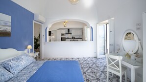 Une chambre à coucher comprenant un lit, un bureau, un miroir et donnant sur la cuisine par une arche.