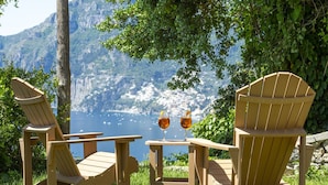 Two wooden Adirondack chairs and a small table with two glasses of amber liquid, set against a backdrop of a mountainous landscape and a body of water. - Punta di Diamante - 200 Steps to Heaven (Praiano)