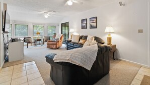 A spacious living room with a large dark sofa, a coffee table, and a dining area with chairs and a table. There are ceiling fans, wall-mounted lamps, and framed pictures on the walls. - Welcome to A Glimpse of the Lake – Your Ideal Lakeside Retreat (Bronston)