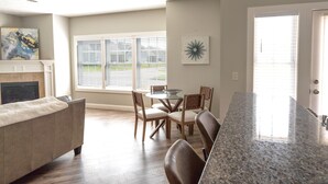 A spacious living room with a fireplace, a dining area with a round table and chairs, and large windows with blinds.