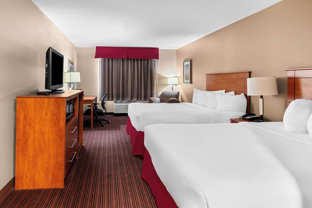 Quarto de hotel com duas camas, uma escrivaninha com televisão, uma cadeira, um abajur e uma janela com cortinas.