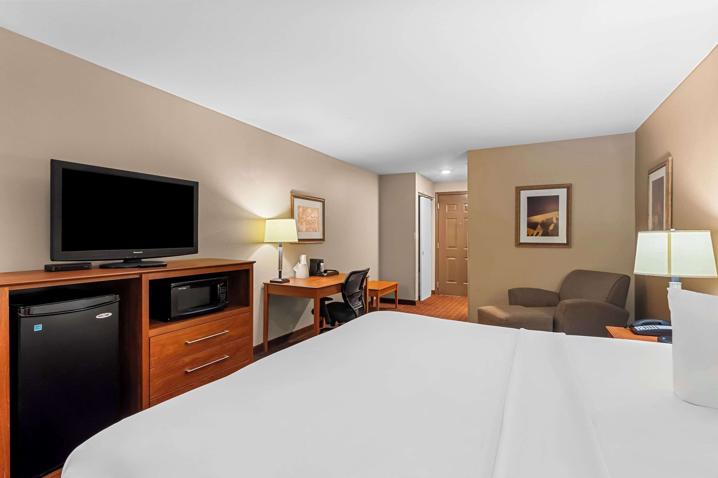 A hotel room with a bed, a desk with a computer, a television, a refrigerator, and two chairs.