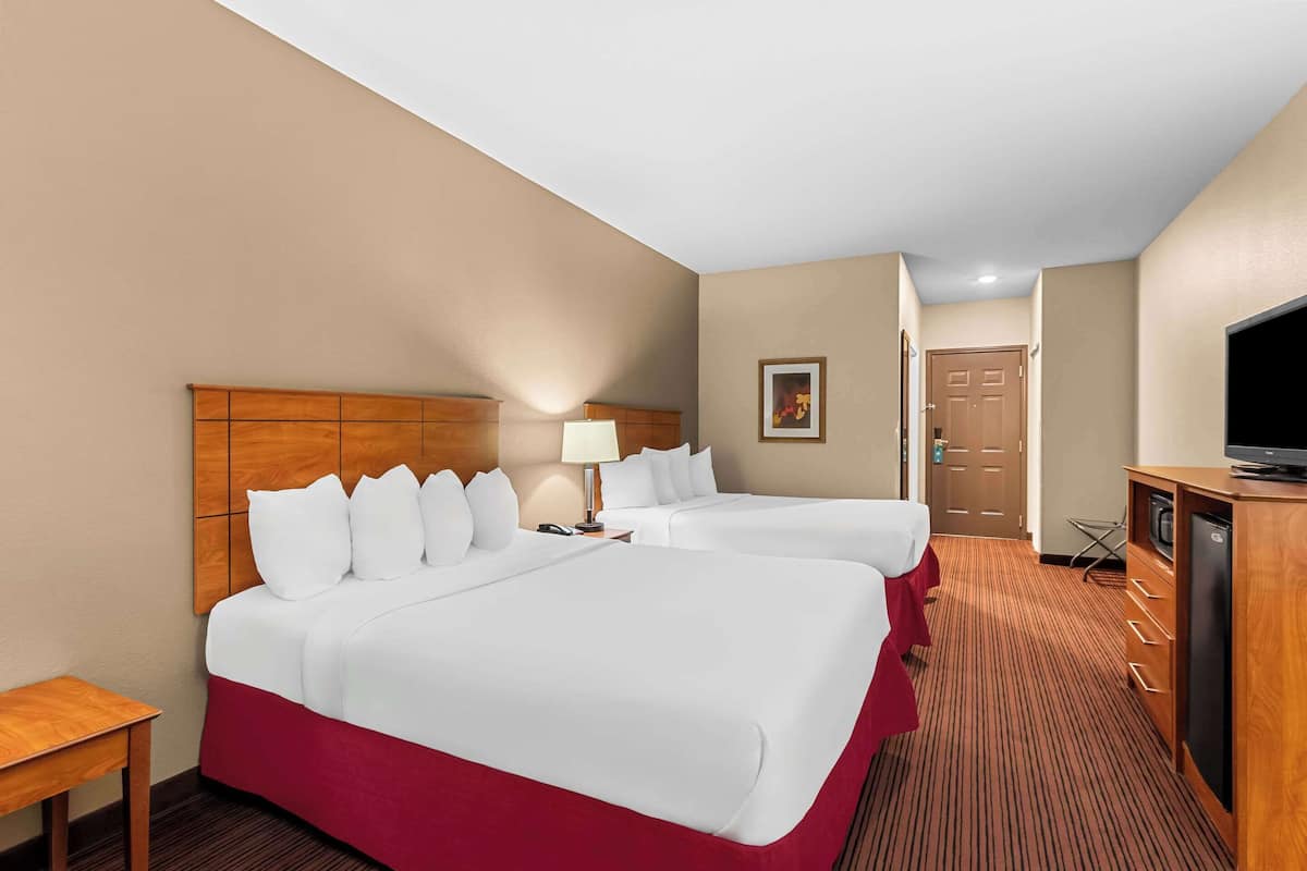 Quarto de hotel com duas camas, cabeceira de madeira, escrivaninha com luminária, televisão e porta ao fundo.