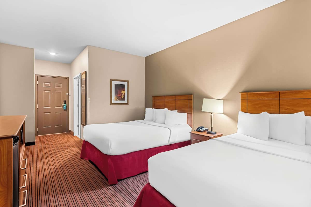 Quarto de hotel com duas camas, uma escrivaninha, uma cadeira, uma mesa de cabeceira com abajur e uma porta.