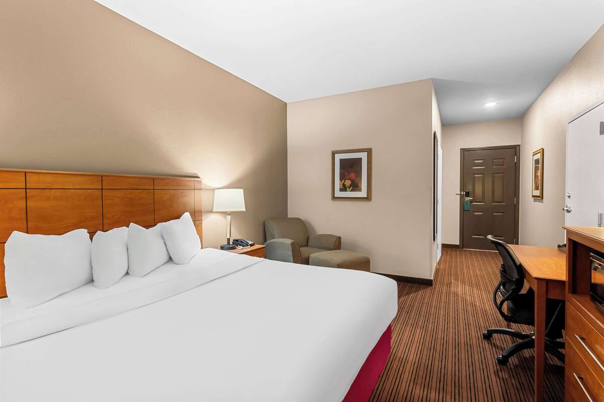 Quarto de hotel com uma cama grande, uma escrivaninha, uma cadeira, um abajur e uma porta.
