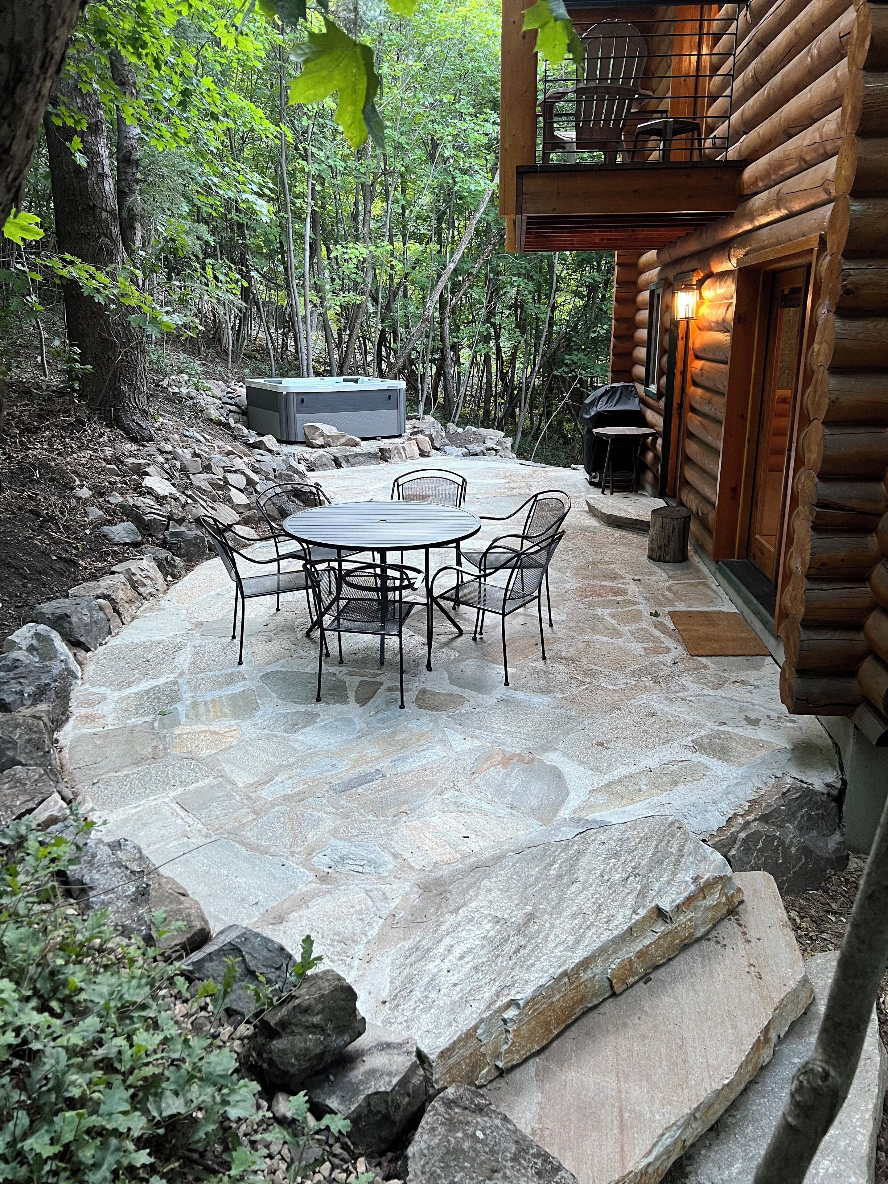 A stone patio with a round table and chairs, surrounded by rocks and greenery, leading to a wooden cabin with a balcony.