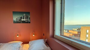 Ein Schlafzimmer mit einem Bett, zwei Nachttischlampen, einem Stadtbildgemälde und einem Fenster mit Blick auf Gebäude und Wasser.