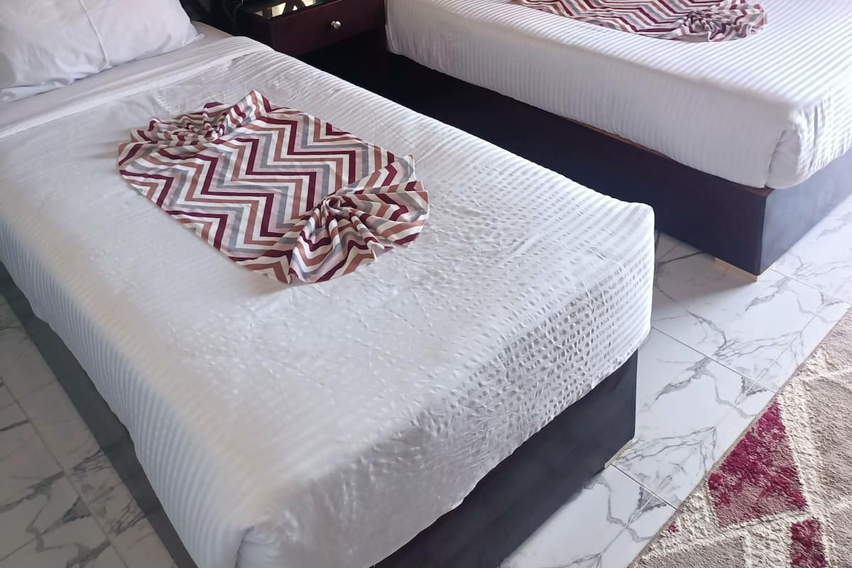 Dos camas con sábanas blancas y mantas estampadas, un cabecero de madera, una mesita de noche con una lámpara y una cortina con diseño.