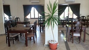 Een ruime eetruimte met meerdere houten tafels en stoelen, een grote plant in pot in het midden en grote ramen met donkere gordijnen.