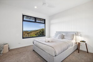 A bedroom with a large window, a bed with striped bedding, a bedside table with a lamp, and a view of a landscape. - Hakea Ridge Coastal Views 2 Decks Family Retreat (Fairhaven)