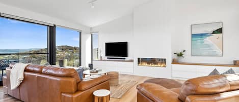 A modern living room with leather sofas, a flat-screen TV, a fireplace, and a large window offering a scenic view.