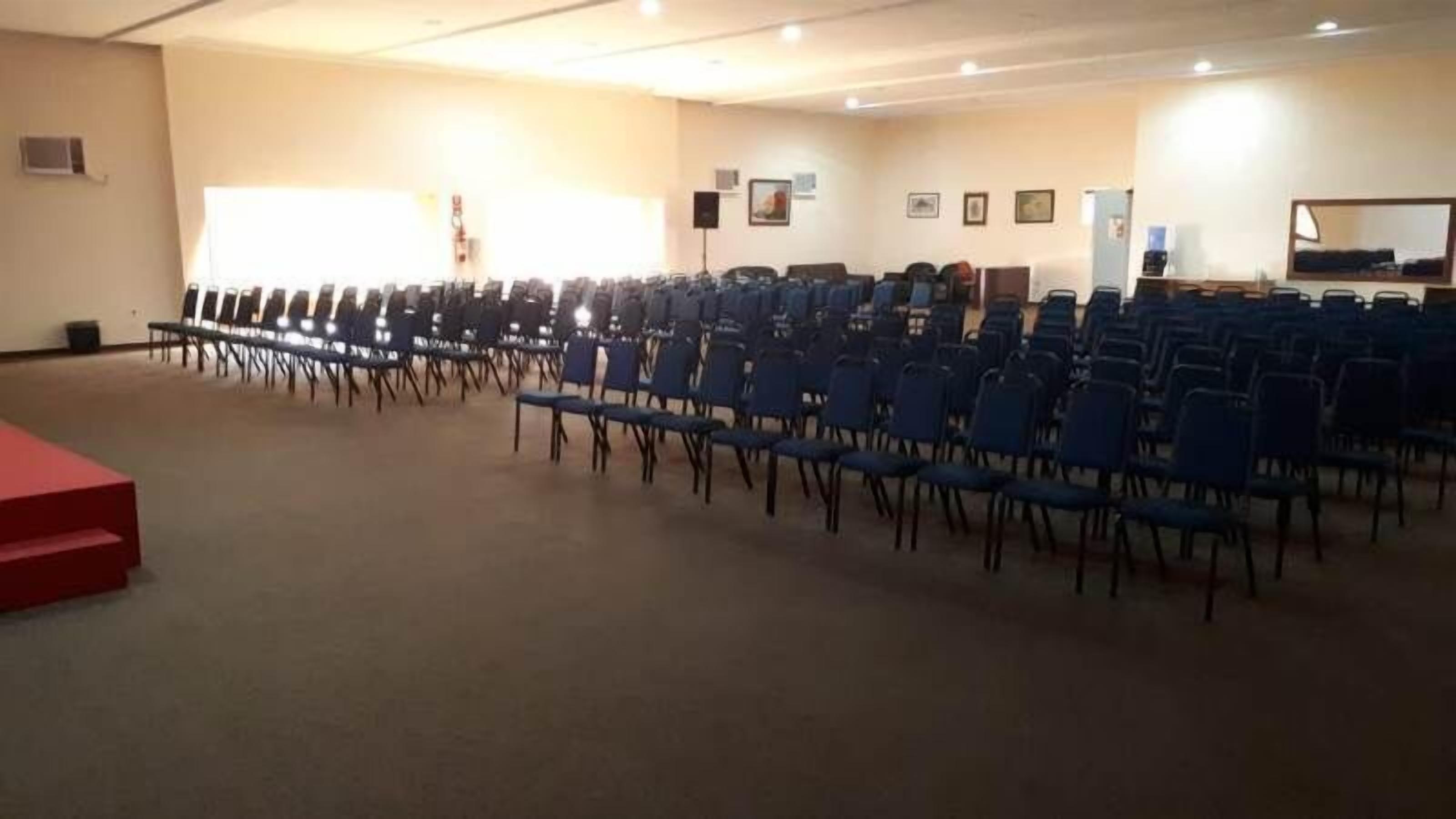 A conference room with rows of blue chairs, a red stage, and a beige carpet.