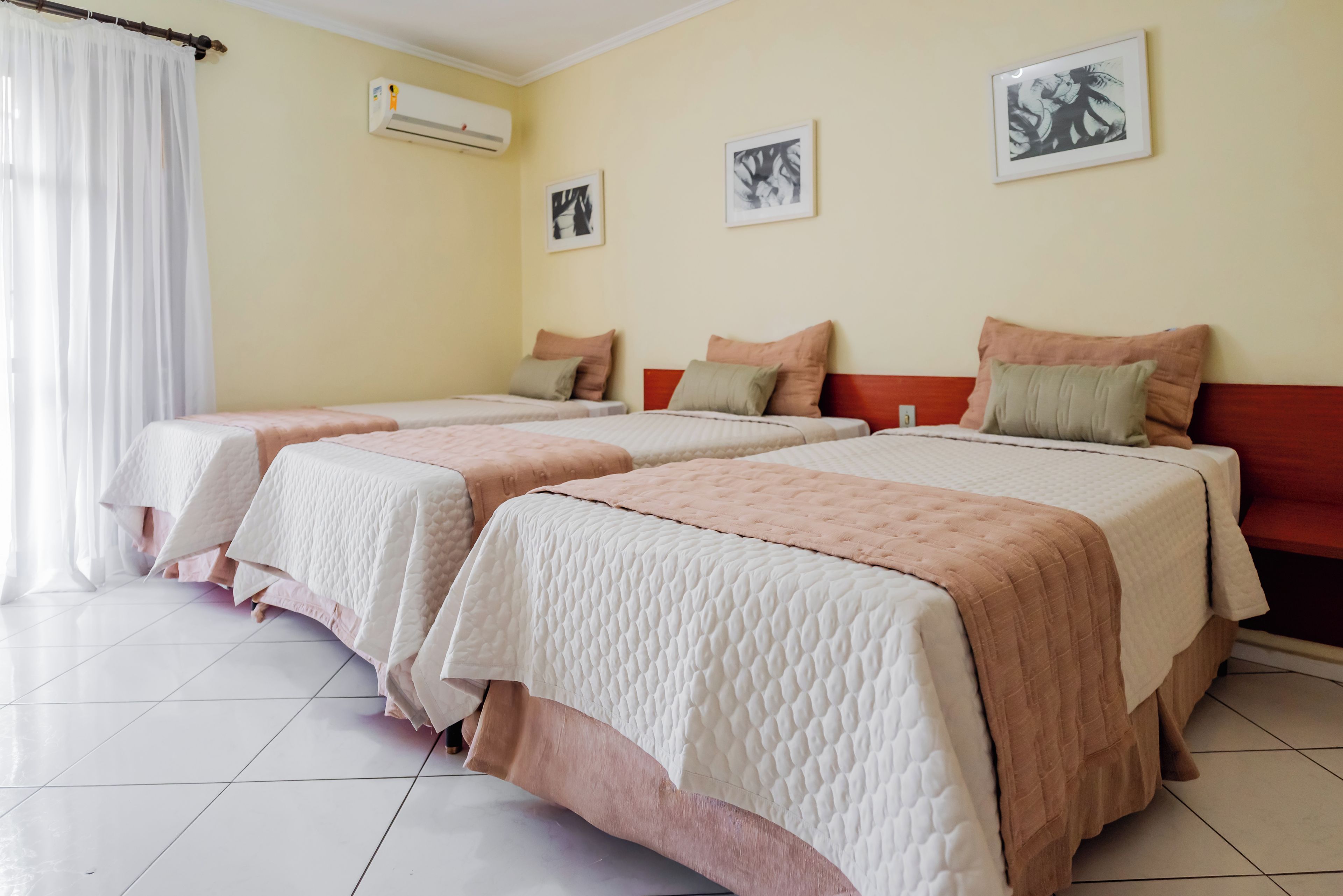 Habitación de hotel con dos camas, una ventana con cortinas blancas, un equipo de aire acondicionado y tres cuadros en la pared.