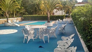Un área junto a la piscina con mobiliario de mimbre blanco, piscina y palmeras.