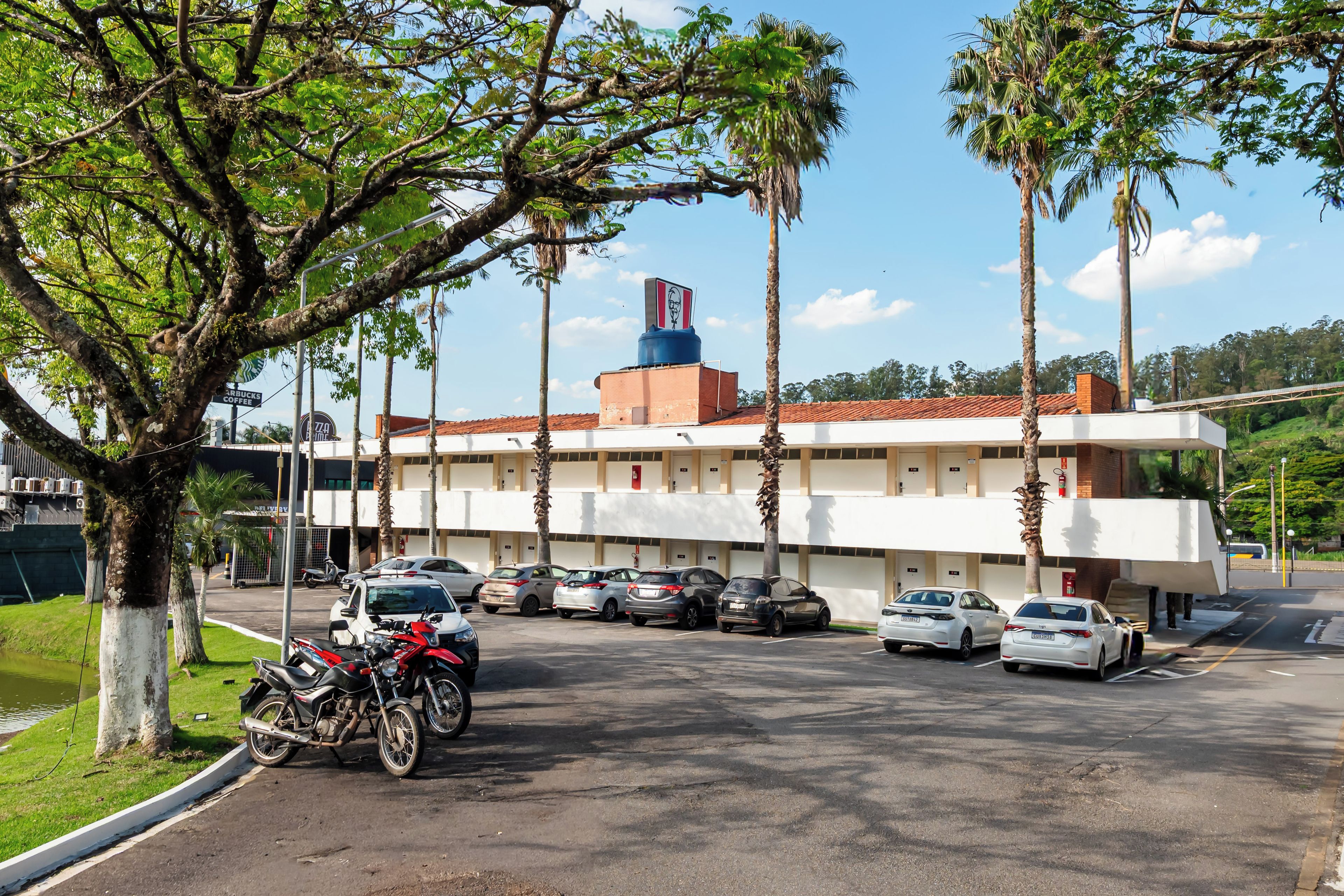 A parking area with several cars and motorcycles, a building with a red roof and a blue water tank, and a tree in the foreground.