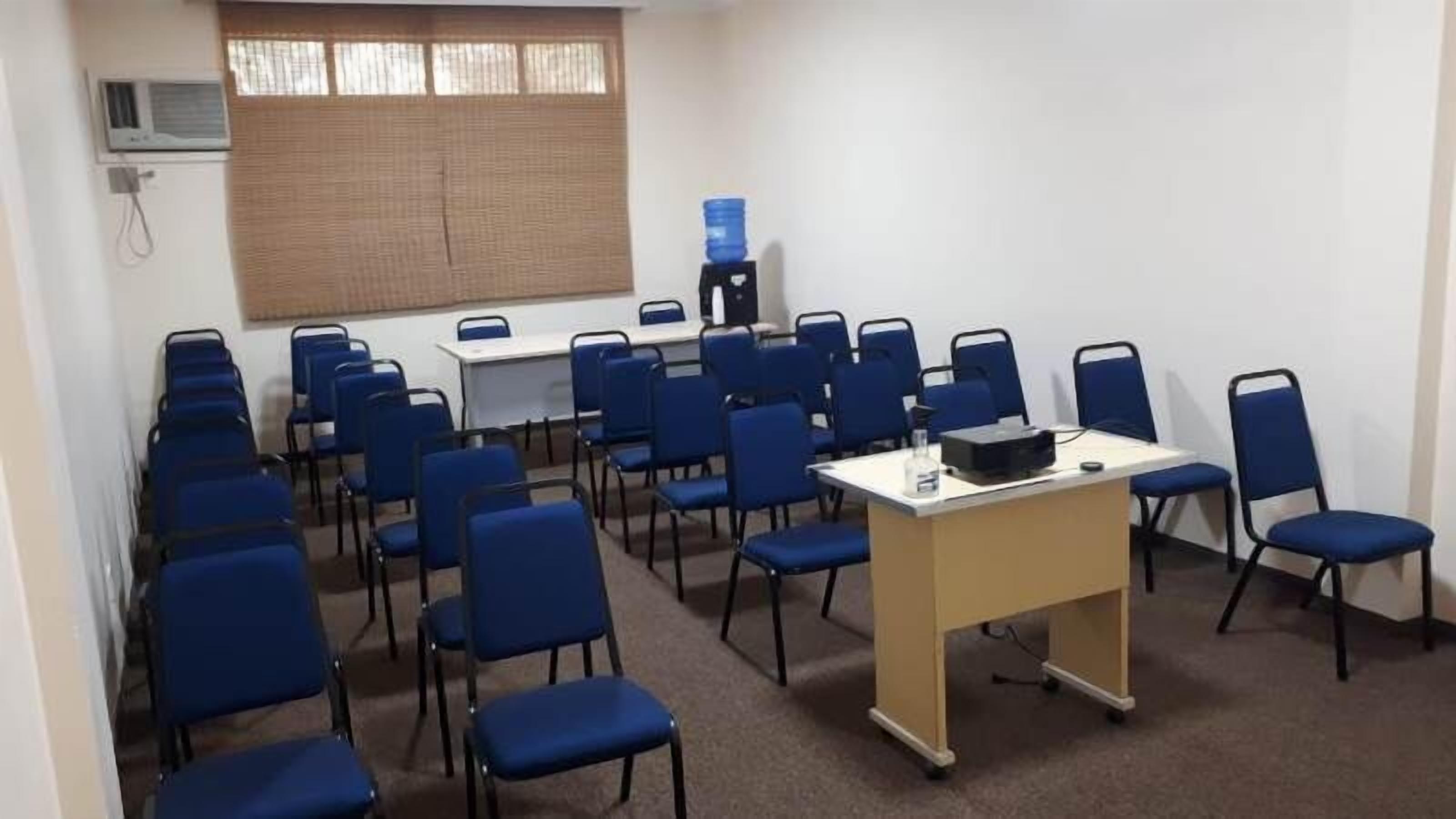 Una sala de conferències amb files de cadires blaves, una taula blanca amb un projector i un dispensador d'aigua.