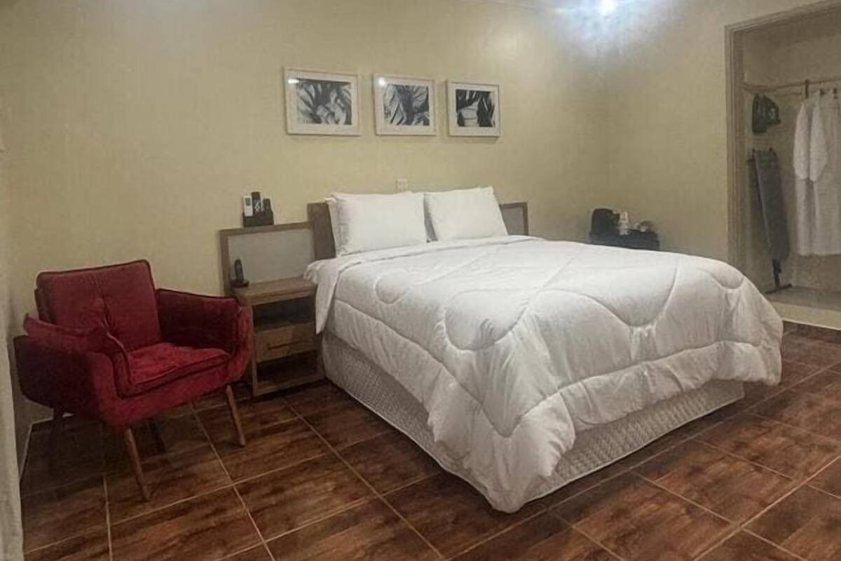 Une chambre à coucher avec un grand lit, un fauteuil rouge, une table de chevet et trois tableaux encadrés accrochés au mur.
