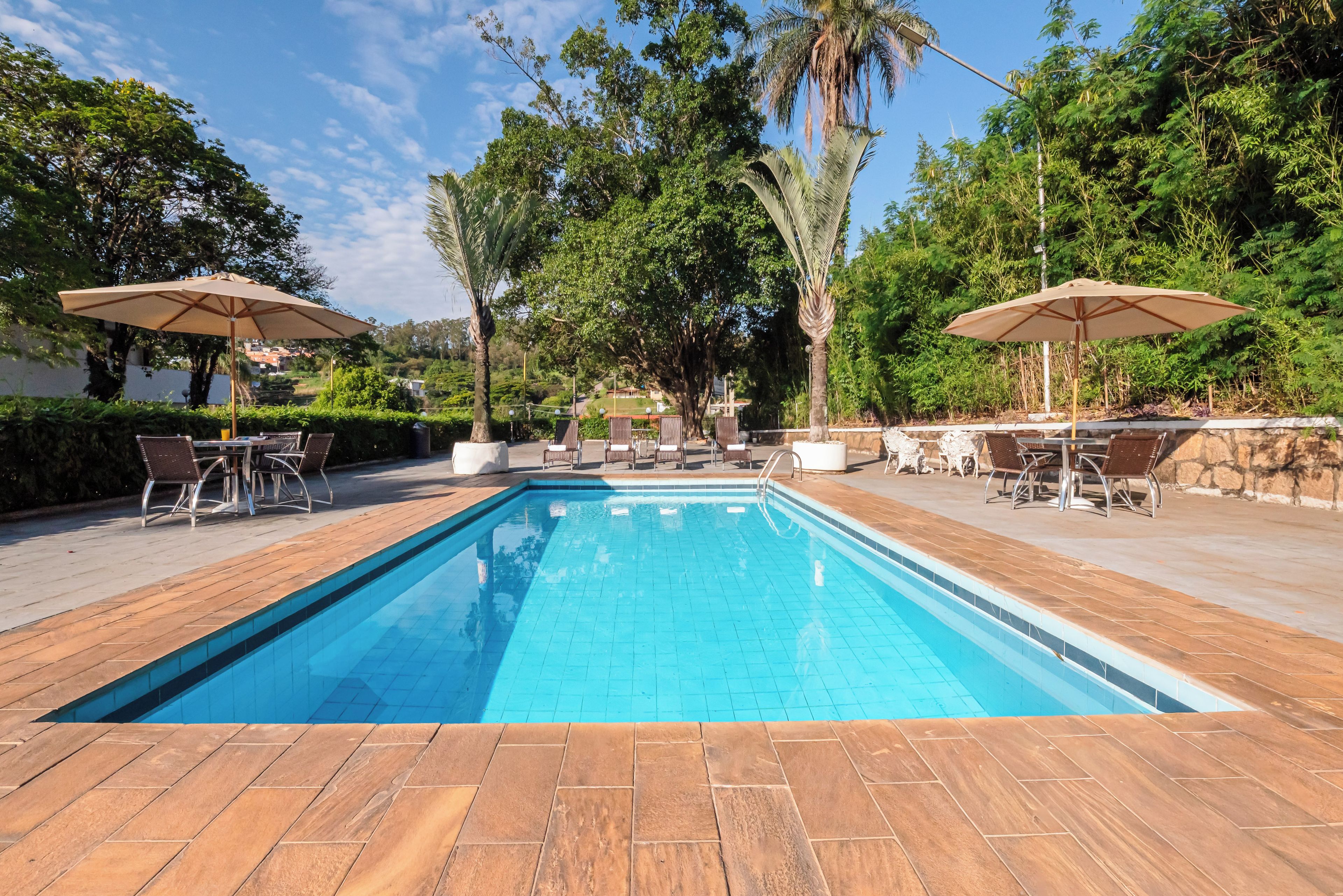 A rectangular swimming pool with a tiled edge, surrounded by wooden decking. There are two outdoor seating areas with umbrellas and chairs, and a stone wall in the background.