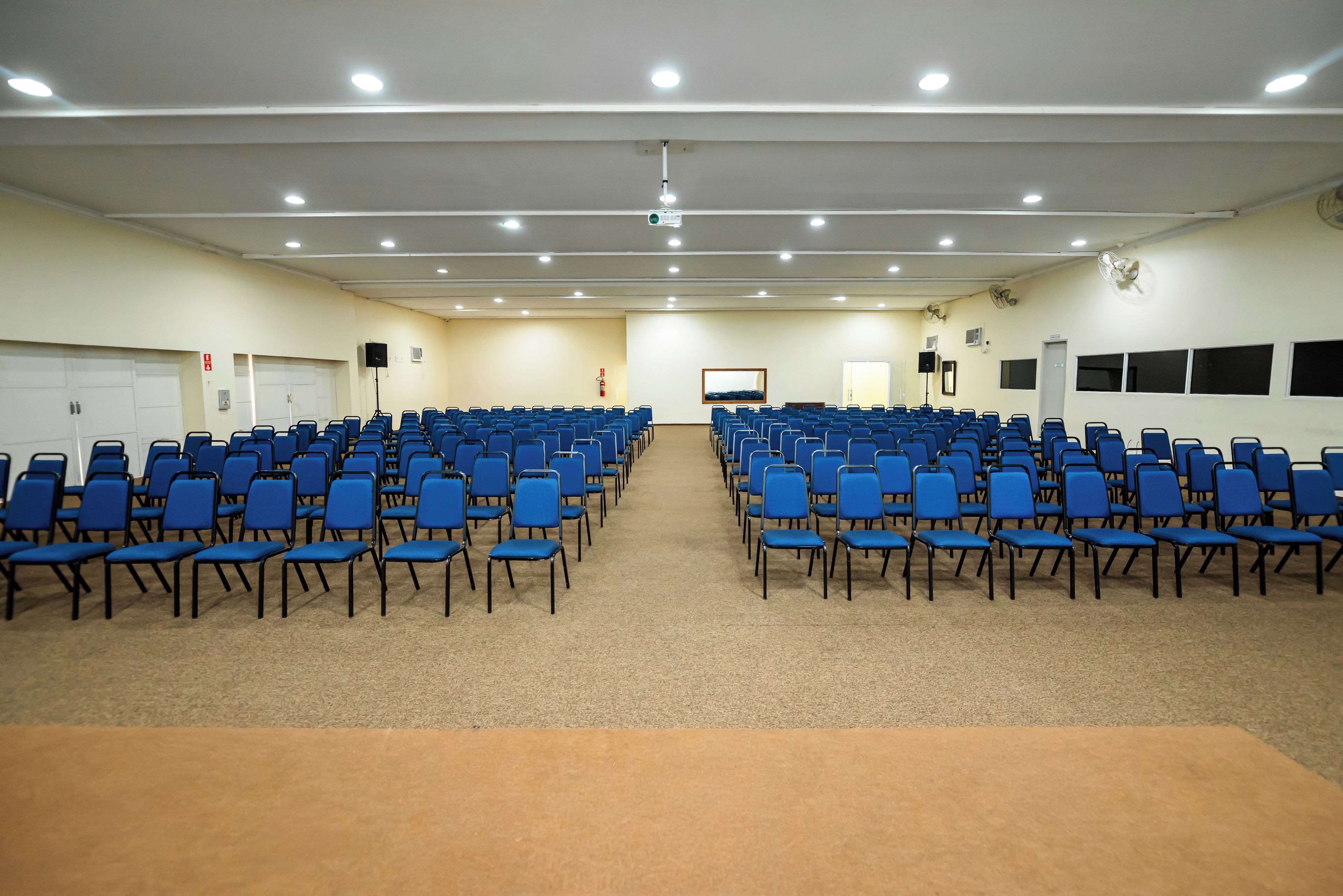 A conference room with rows of blue chairs, a carpeted floor, and a white ceiling with recessed lighting.