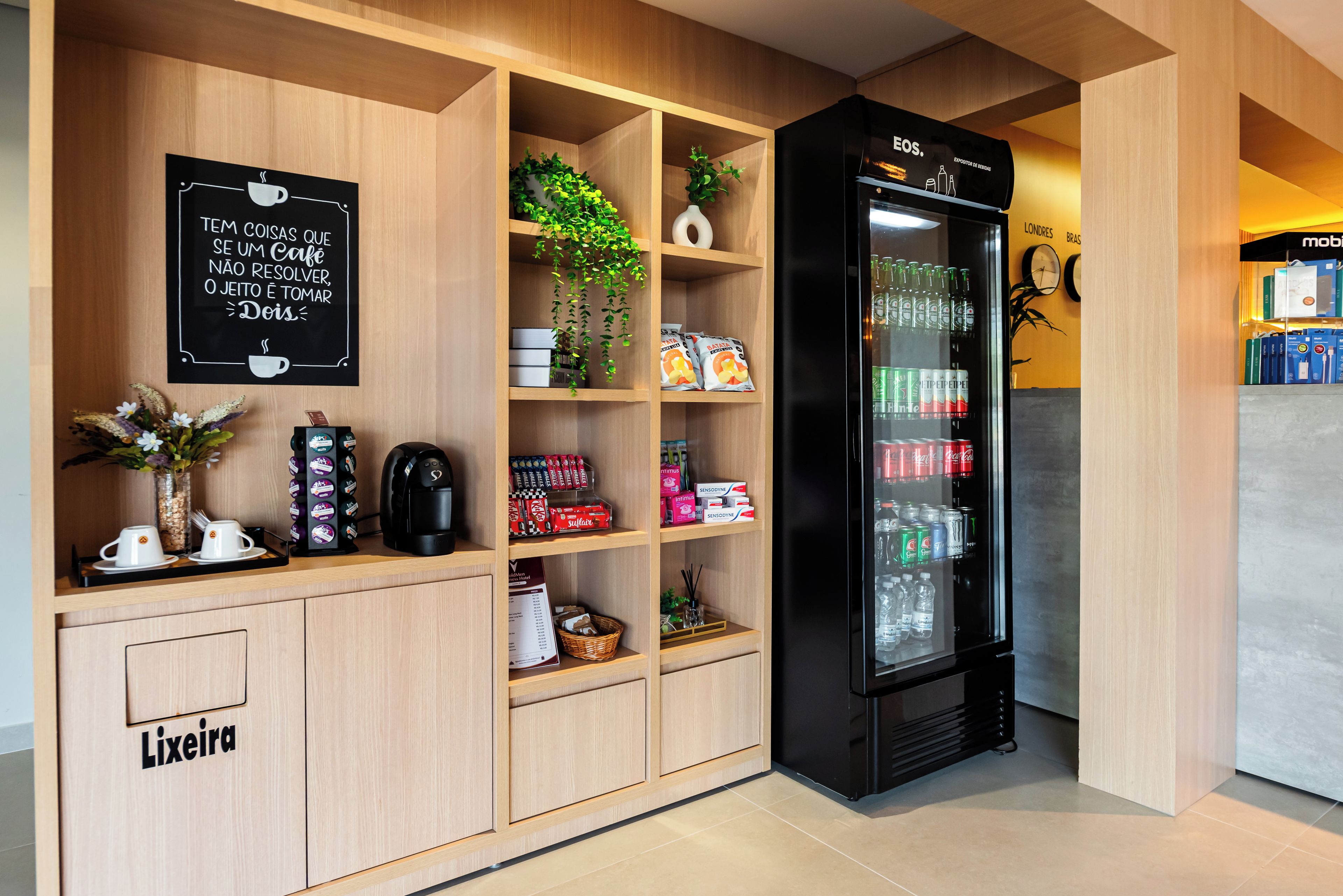 Un minibár de hotel con una cafetera, tazas para té y café, una planta, aperitivos y un refrigerador lleno de bebidas.