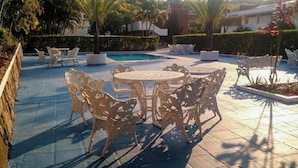 Zona de piscina al aire libre con sillas y mesas blancas ornamentadas, rodeada de palmeras y un seto.