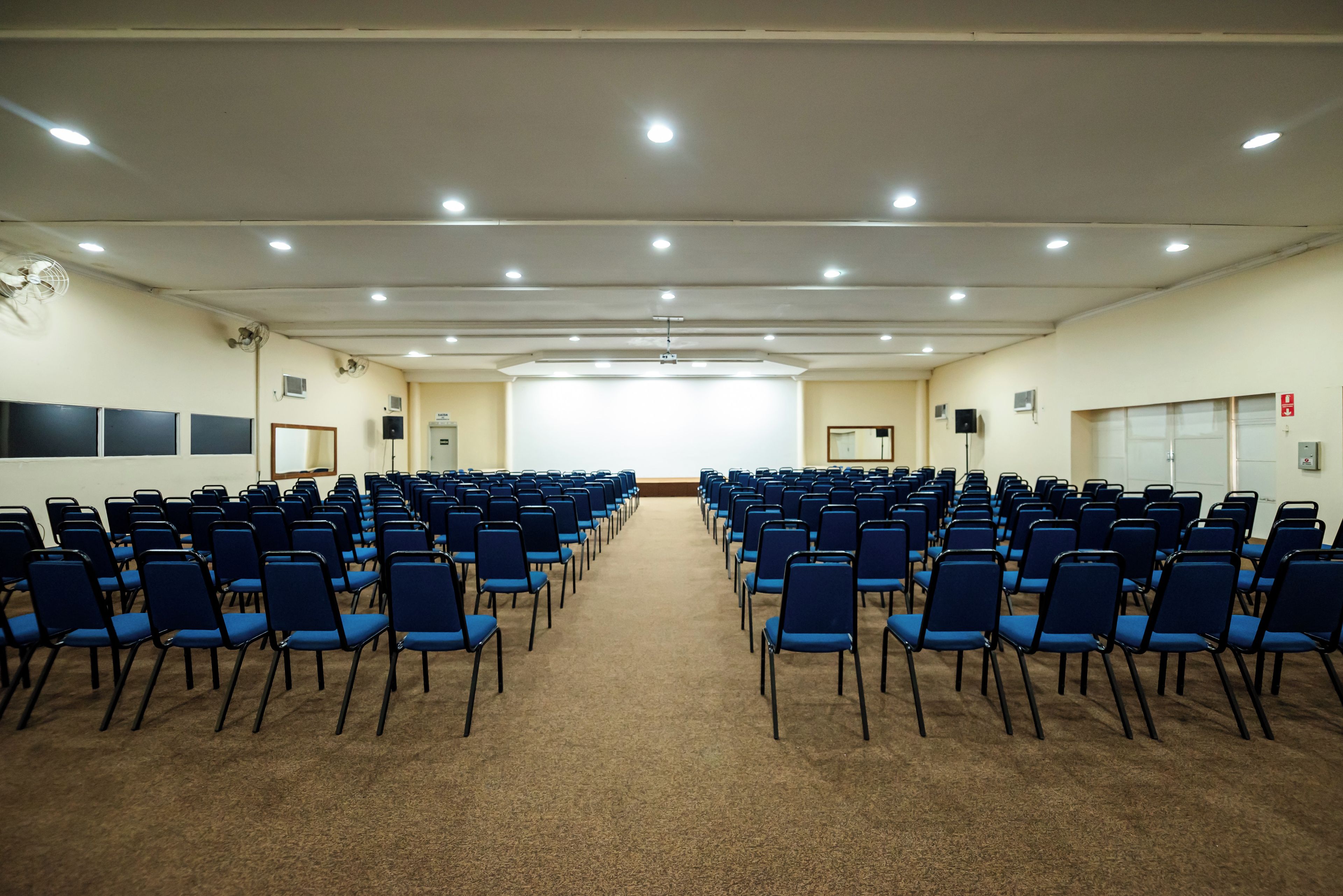 A conference room with rows of blue chairs, carpeted floor, and ceiling lights.