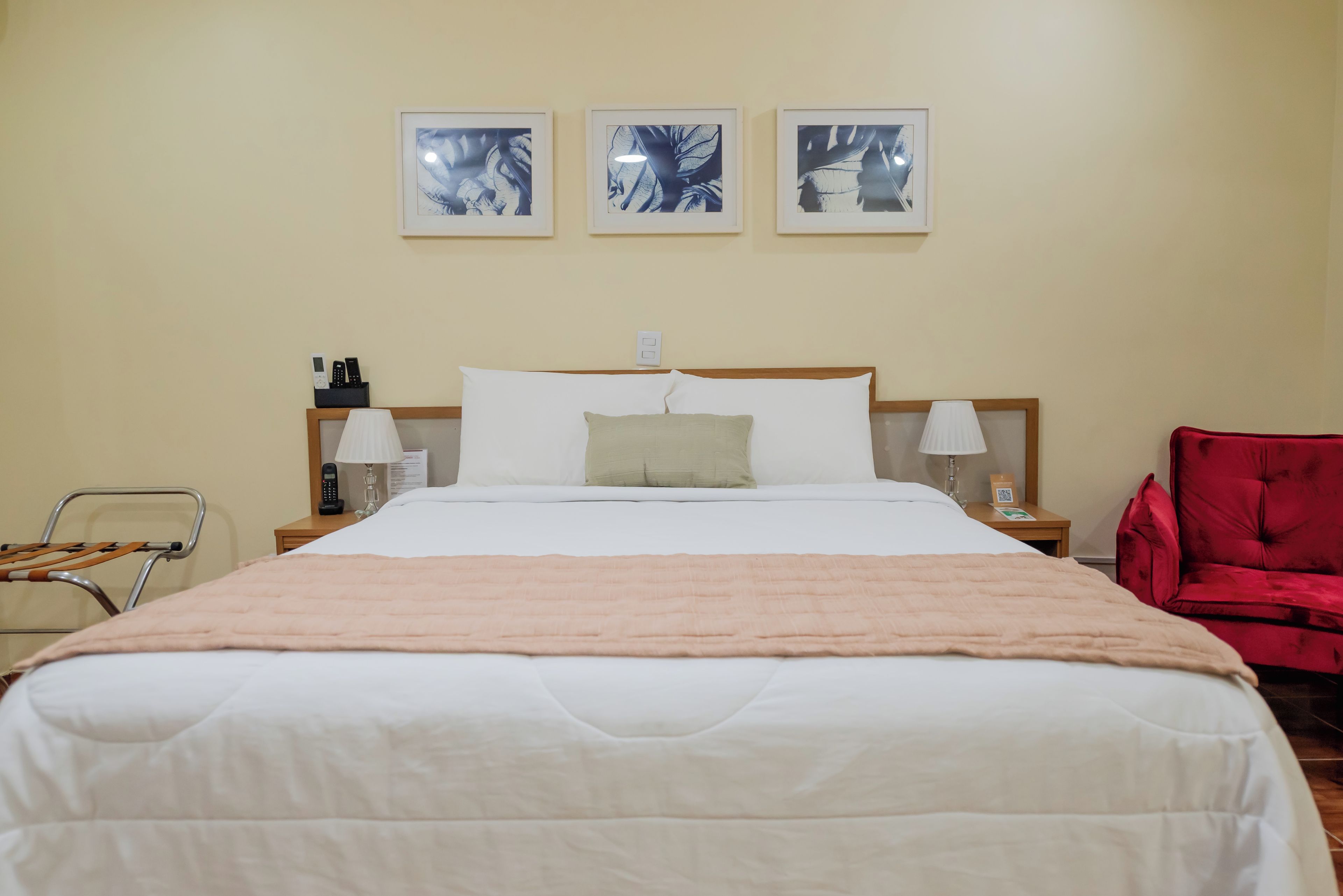 A hotel room with a large bed, a red chair, a wooden nightstand, a lamp, and three framed pictures on the wall.